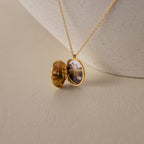 The Eliza Oval Locket Necklace opens to display a small photo inside, resting on a textured light surface—an ideal sentimental gift for preserving cherished memories.