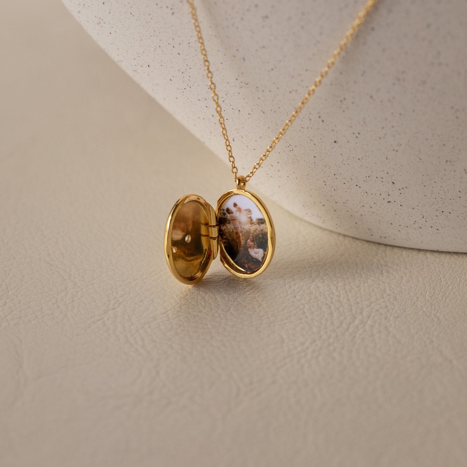 The Eliza Oval Locket Necklace opens to display a small photo inside, resting on a textured light surface—an ideal sentimental gift for preserving cherished memories.