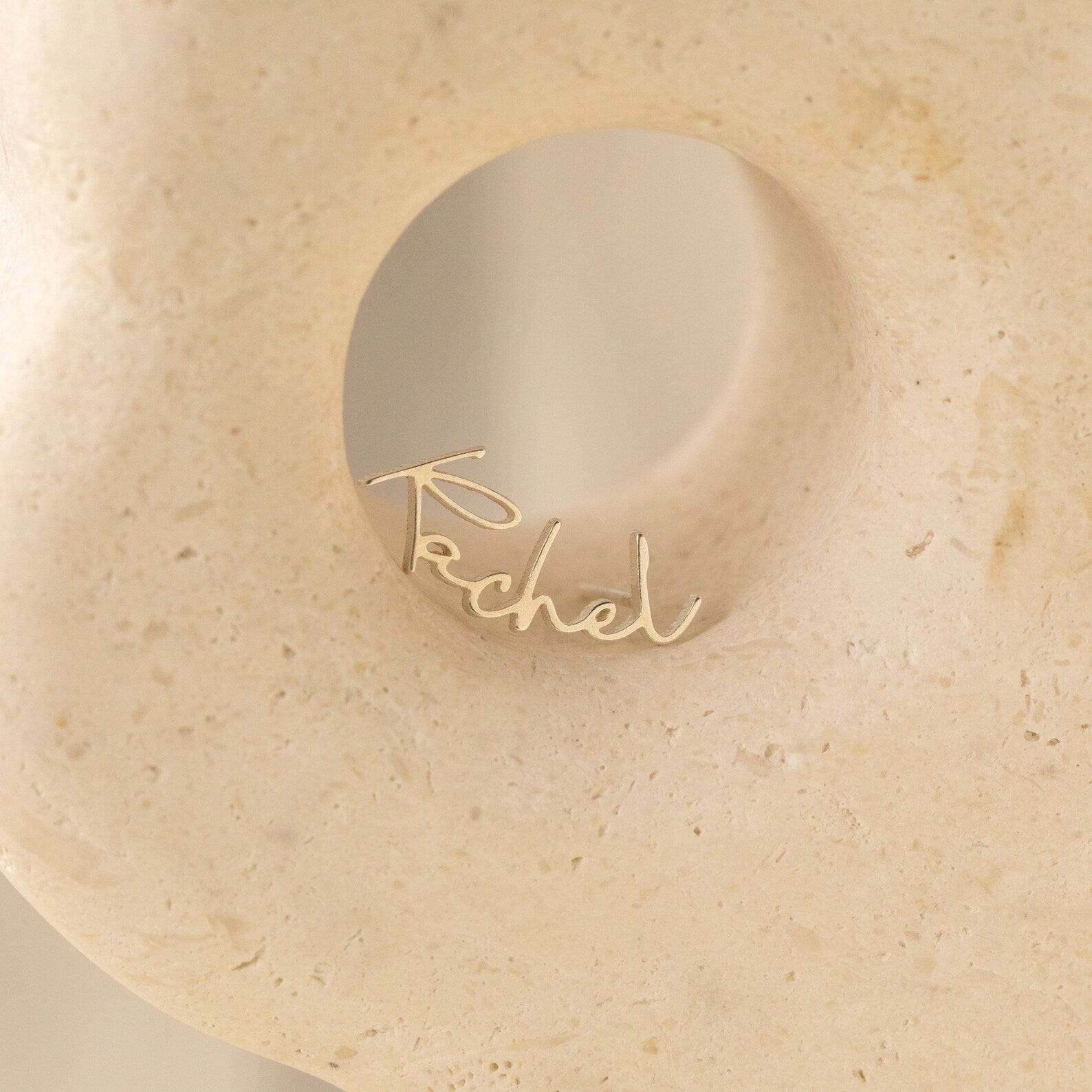 The Summer Name Earrings, gold wire spelling out Rachel in cursive, are displayed in a circular hollow of a smooth, pale stone sculpture, highlighting the charm of personalized jewelry.