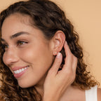 A woman with curly hair smiles and touches her Opal Heart Studs earring, radiating minimalistic glam against a beige background.