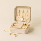 The Square Velvet Jewelry Case in beige velvet is open, displaying gold necklaces, rings, and earrings neatly organized inside and beside it.