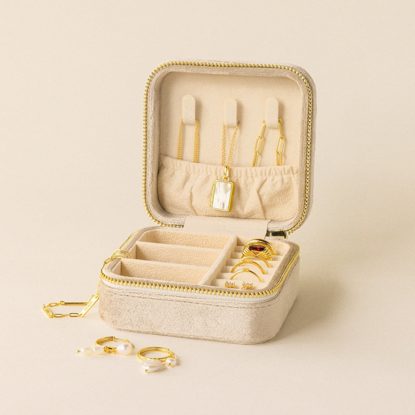 The Square Velvet Jewelry Case in beige velvet is open, displaying gold necklaces, rings, and earrings neatly organized inside and beside it.