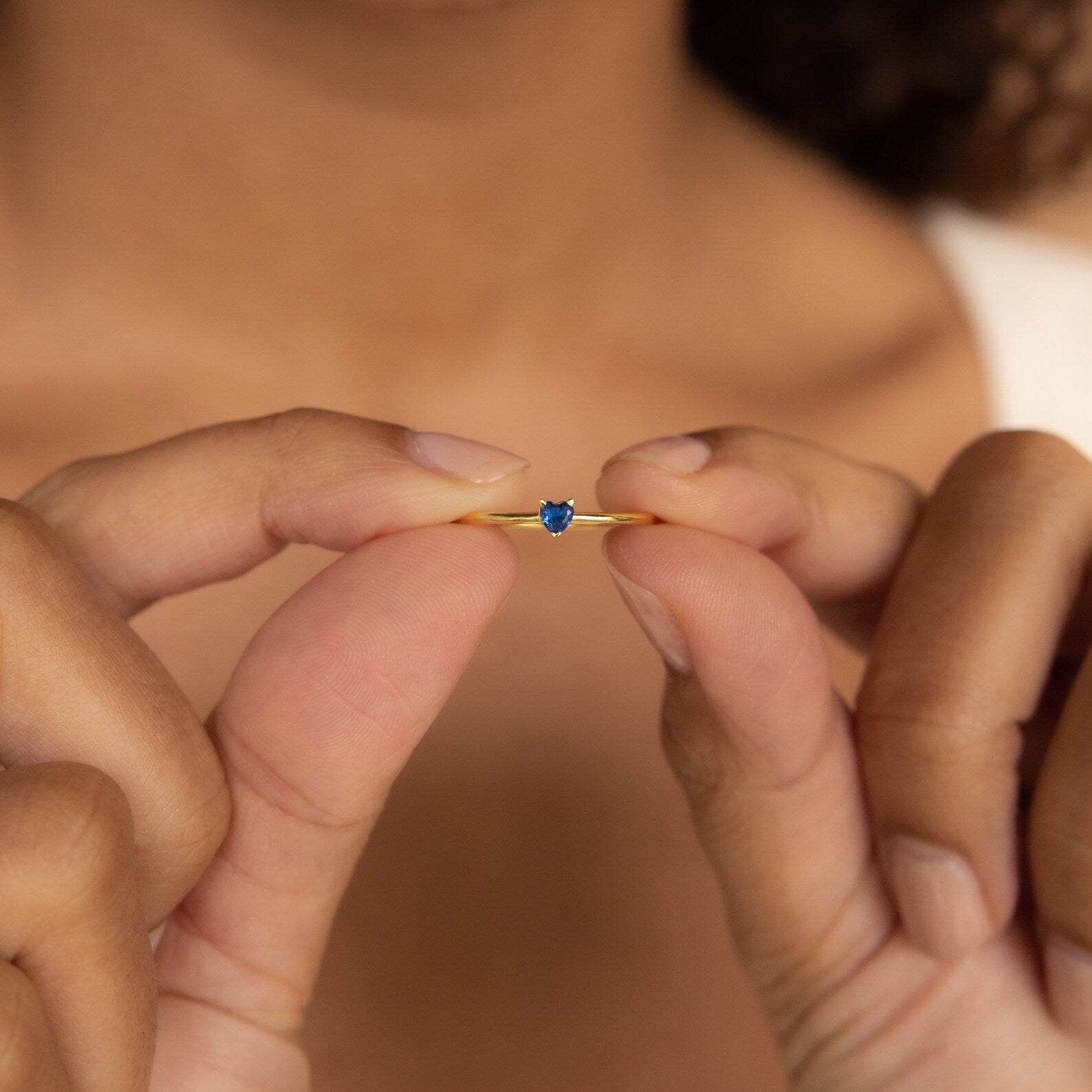 A person holds the Tiny Heart Birthstone Ring—a delicate gold stacking ring featuring a small blue gemstone—between their fingers.