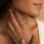 A woman gently touches her neck, wearing the Tiny Heart Birthstone Ring—a delicate gold stacking ring with a small blue stone—on her finger.