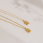 On a light surface with soft floral shadows, the Ribbed Flower Tag Necklace exudes timeless charm and subtle elegance, evoking vintage-inspired style.