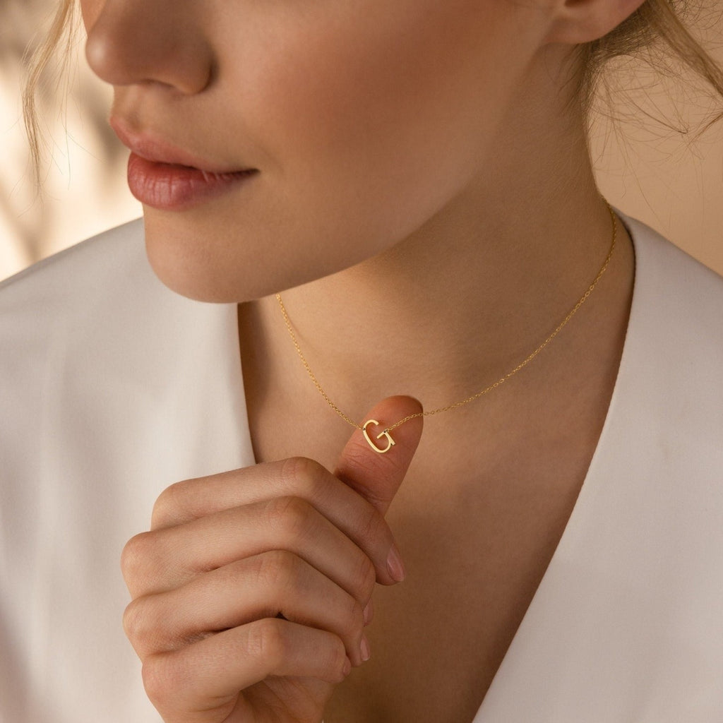 A woman gently holds her Mini Sideways Initial Necklace, featuring a gold letter G pendant—a beautiful personalized accessory.