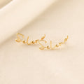 Two Summer Name Earrings rest on soft, cream-colored fabric. Each earring features elegant cursive script, spelling Selena—a stunning example of personalized jewelry.