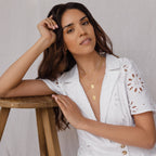 A woman in a white eyelet dress poses by a wooden stool, accessorized with layered gold necklaces, including the Birth Flower Arch Necklace, and wears subtle makeup.