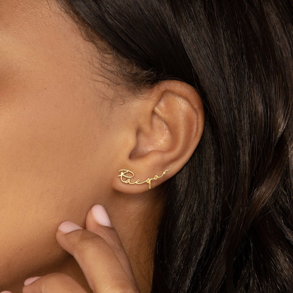 A woman wears the Hallie Name Earrings, a gold stud spelling "Raegan" in cursive, as she touches her face—showcasing personalized jewelry at its finest.