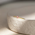 The Tiny Heart Birthstone Ring features a delicate pink gemstone set in a gold heart, displayed on a textured off-white surface—ideal as a romantic gift or unique stacking ring for celebrating a special birth month.