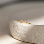 The Tiny Heart Birthstone Ring features a delicate pink gemstone set in a gold heart, displayed on a textured off-white surface—ideal as a romantic gift or unique stacking ring for celebrating a special birth month.
