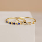 The Space Birthstone Ring, a delicate gold band featuring small blue stones, rests elegantly on a white surface.