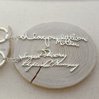 Showcasing two silver necklaces with cursive handwriting pendants on wood; one says "I love you little one Mother." These unique pieces pair perfectly with our Custom Handwriting Keychain—ideal for heartfelt, personalized gifts.