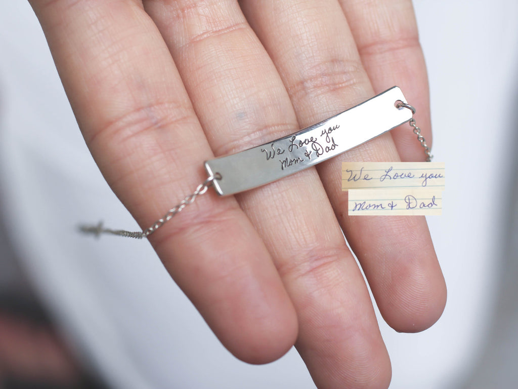 A hand holds a Handwriting Bar Bracelet, a silver bracelet custom-engraved with “We Love you Mom & Dad” in your own handwriting.