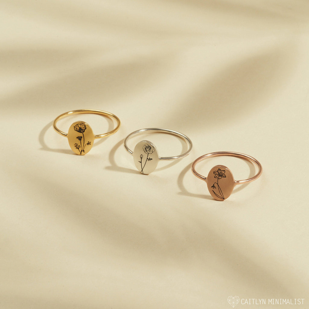 The Birth Flower Oval Ring features minimalist gold, silver, and rose gold bands, each engraved with a birth month flower—a stylish choice for stacking or gifting. Shown on a beige background.