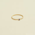 The Twisted Birthstone Ring features a delicate gold twist design with a small purple gemstone, perfect for adding color to your stack, displayed on a light beige background.