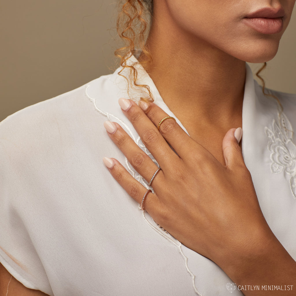 A woman in a white blouse displays her hand adorned with three delicate rings, including the Lea Twisted Ring and polished bands on her fingers.