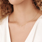 A woman wears the Emery Necklace in Sterling Silver—a minimal piece with a delicate chain and tiny pendant—paired with a white top. This necklace makes a perfect holiday gift.