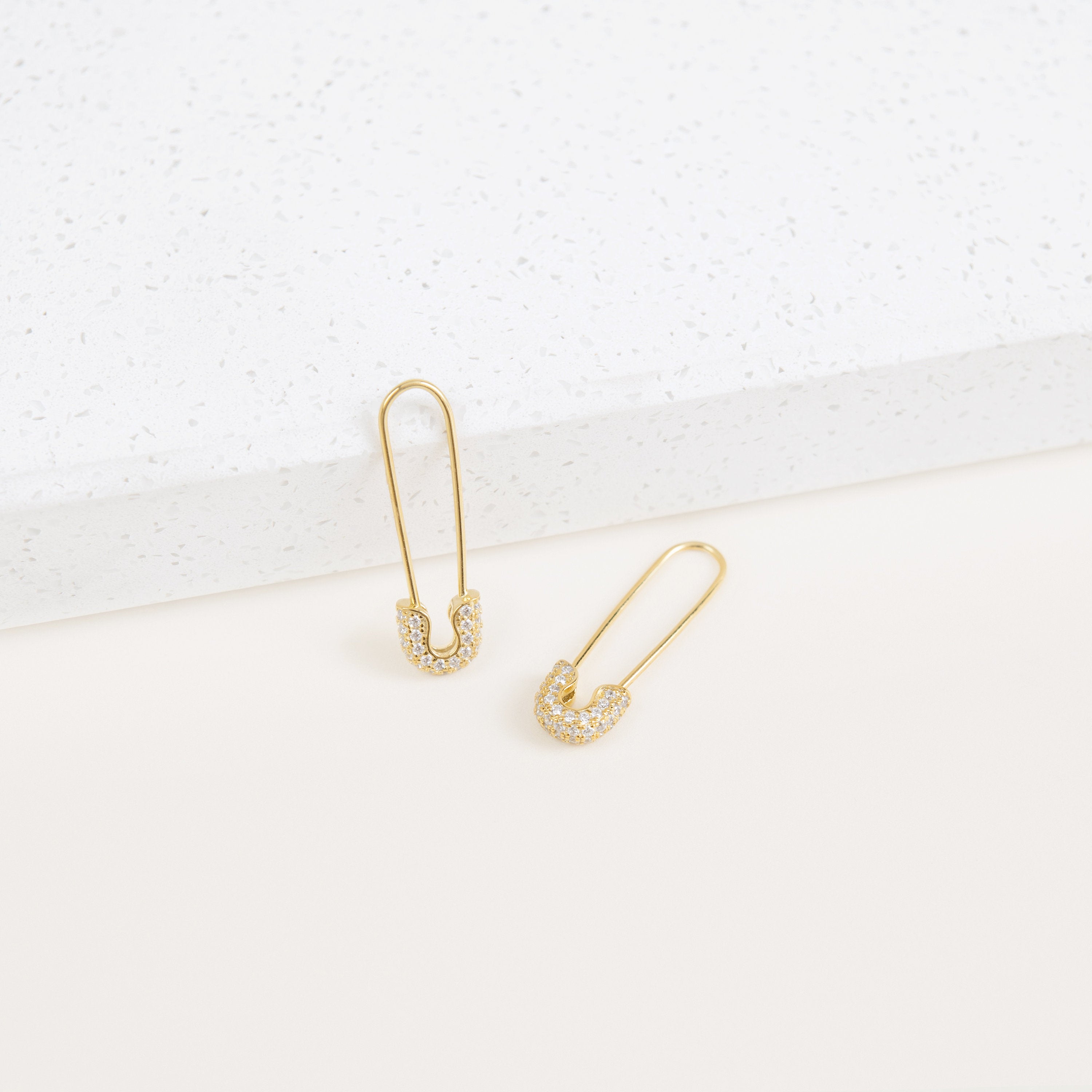 The Pave Safety Pin Earrings, accented with gold and diamonds, showcase their unique safety pin design as they rest elegantly on a white textured surface.