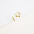 The Remi Twist Hoops, a pair of gold earrings, are displayed on a white textured surface with a minimalist background—perfect for fans of hoop earrings and minimal jewelry.