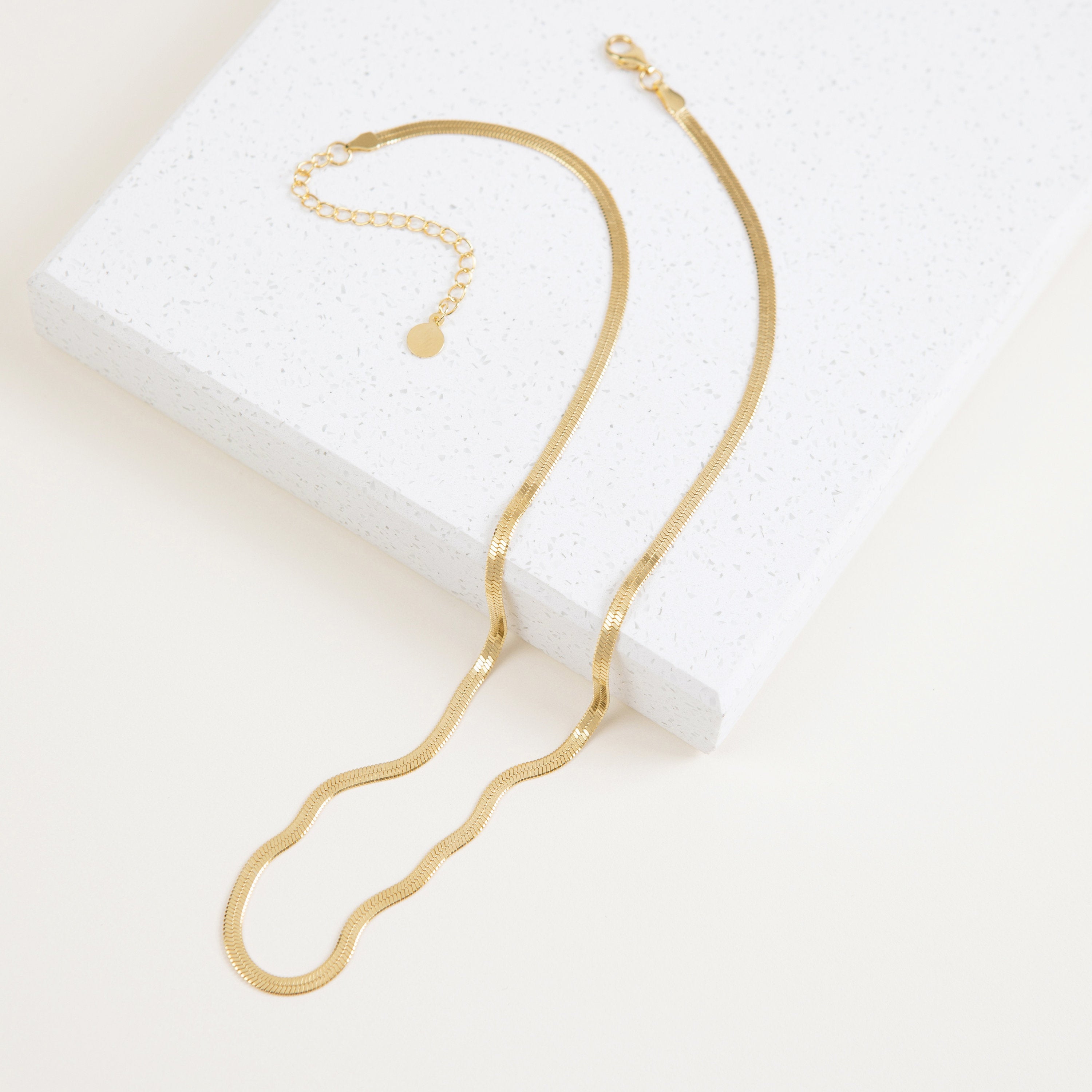 A close-up view of a gold herringbone necklace resting on a terrazzo display block.