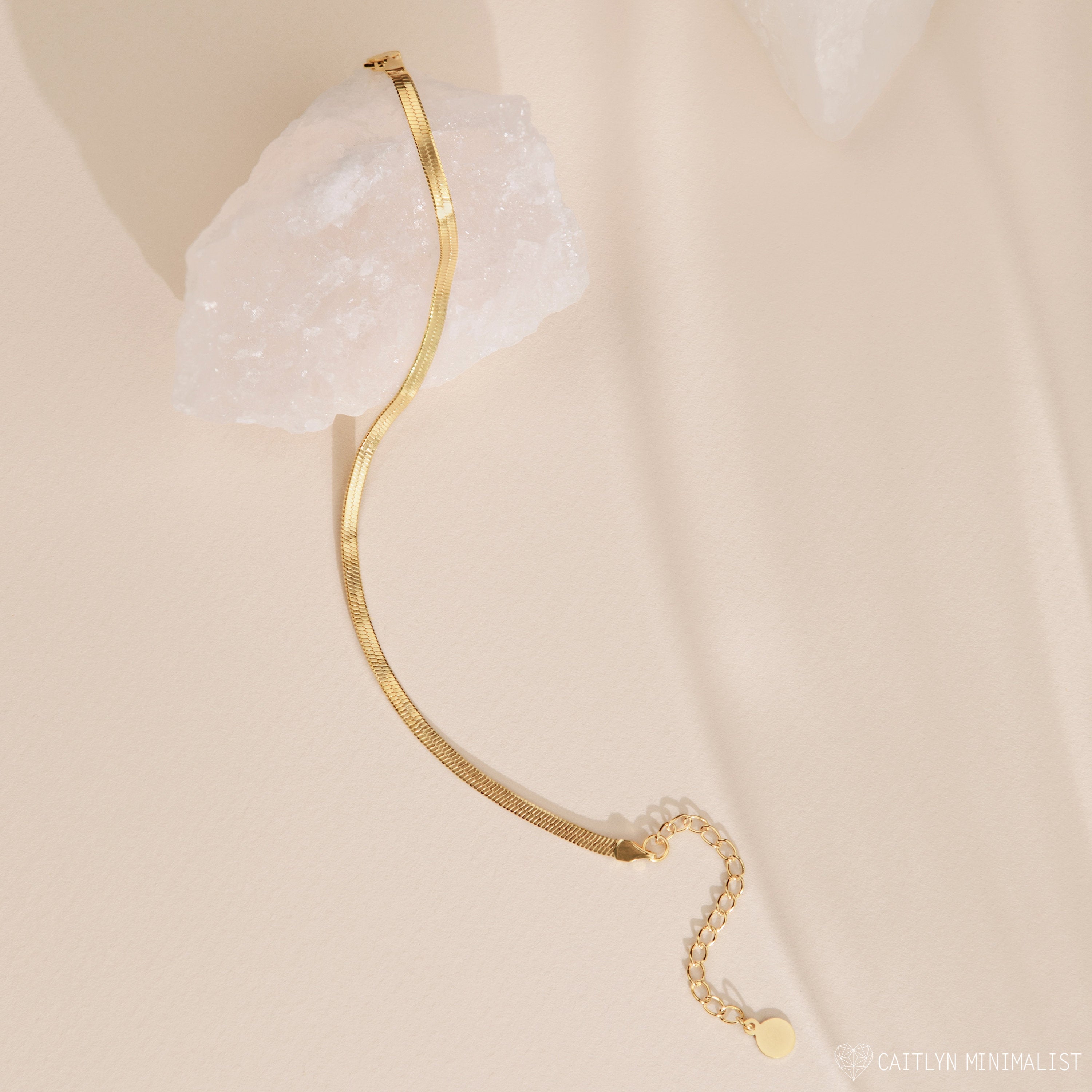 The Herringbone Bracelet features a sleek gold chain design, elegantly displayed over a white crystal on a light beige background.