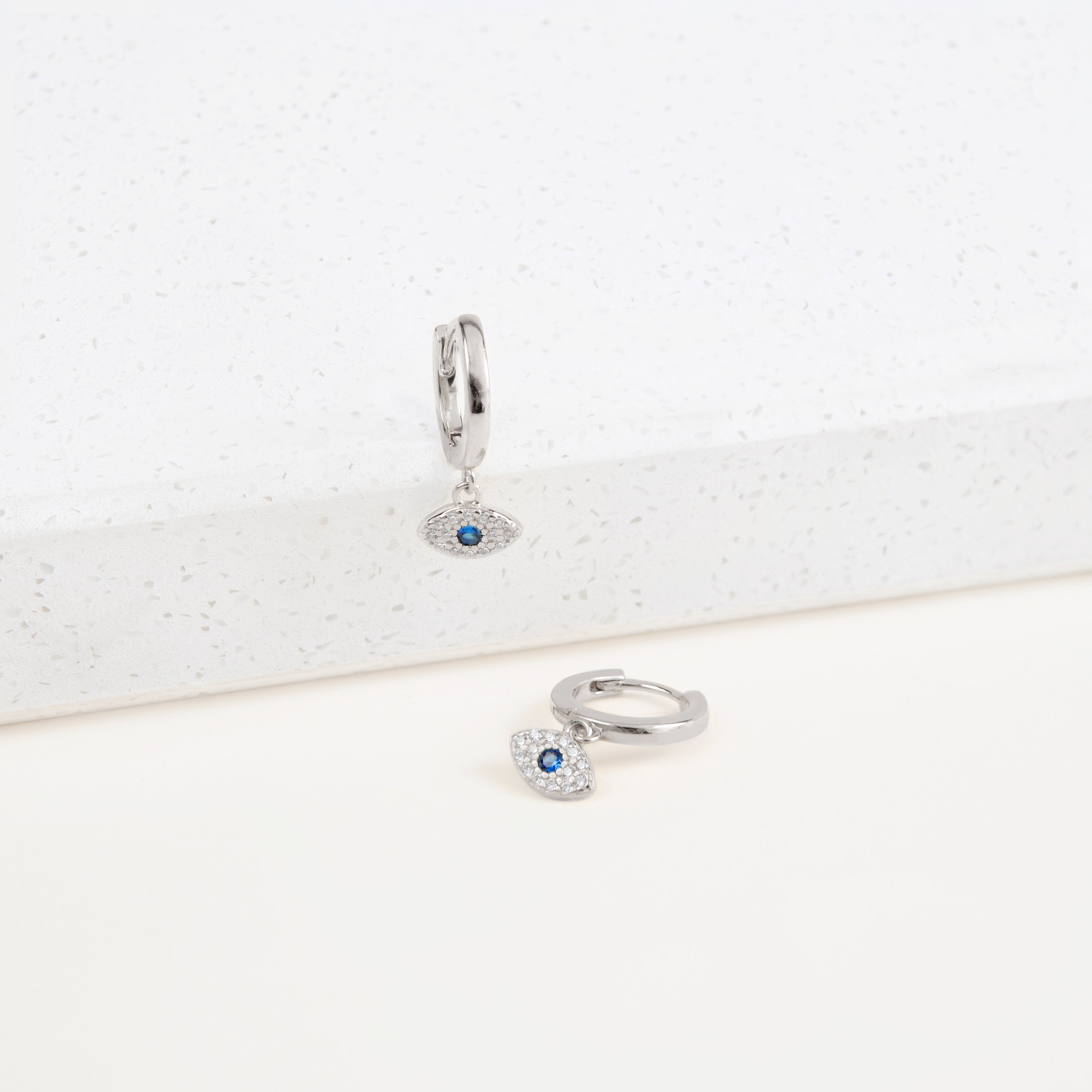 The Iris Huggies are silver huggie earrings featuring blue and white evil eye charms, beautifully displayed on a white surface—ideal as stylish protection jewelry.