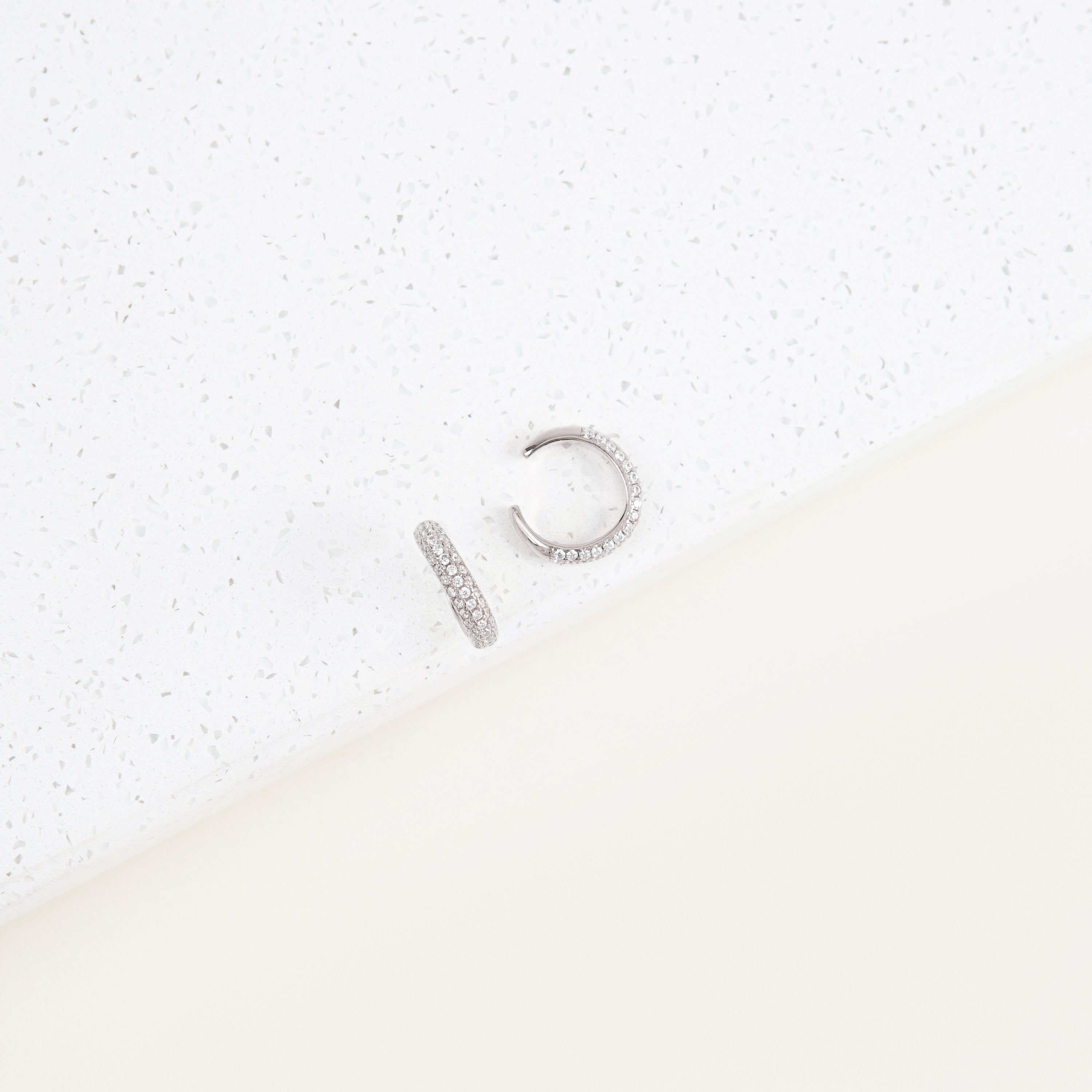The Mia Pave Cuffs—sparkling silver hoops accented with clear stones—rest elegantly on a white speckled surface.