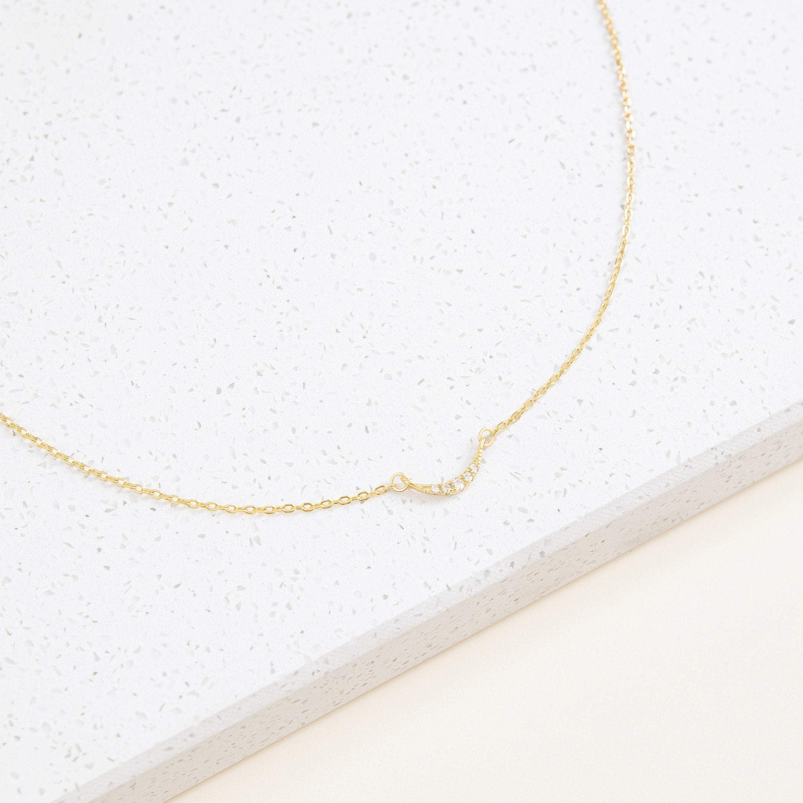 The Lucia Pave Necklace is a delicate gold chain with a small crescent charm pendant adorned with tiny crystals, beautifully displayed on a white surface.