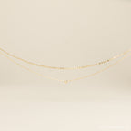 The Zina Necklace, a delicate gold chain with a single bead, is displayed on a neutral background.