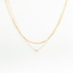The Zina Necklace features two delicate gold chains—a double-layered design with a small gold bead and a simple pendant—displayed on a white background.