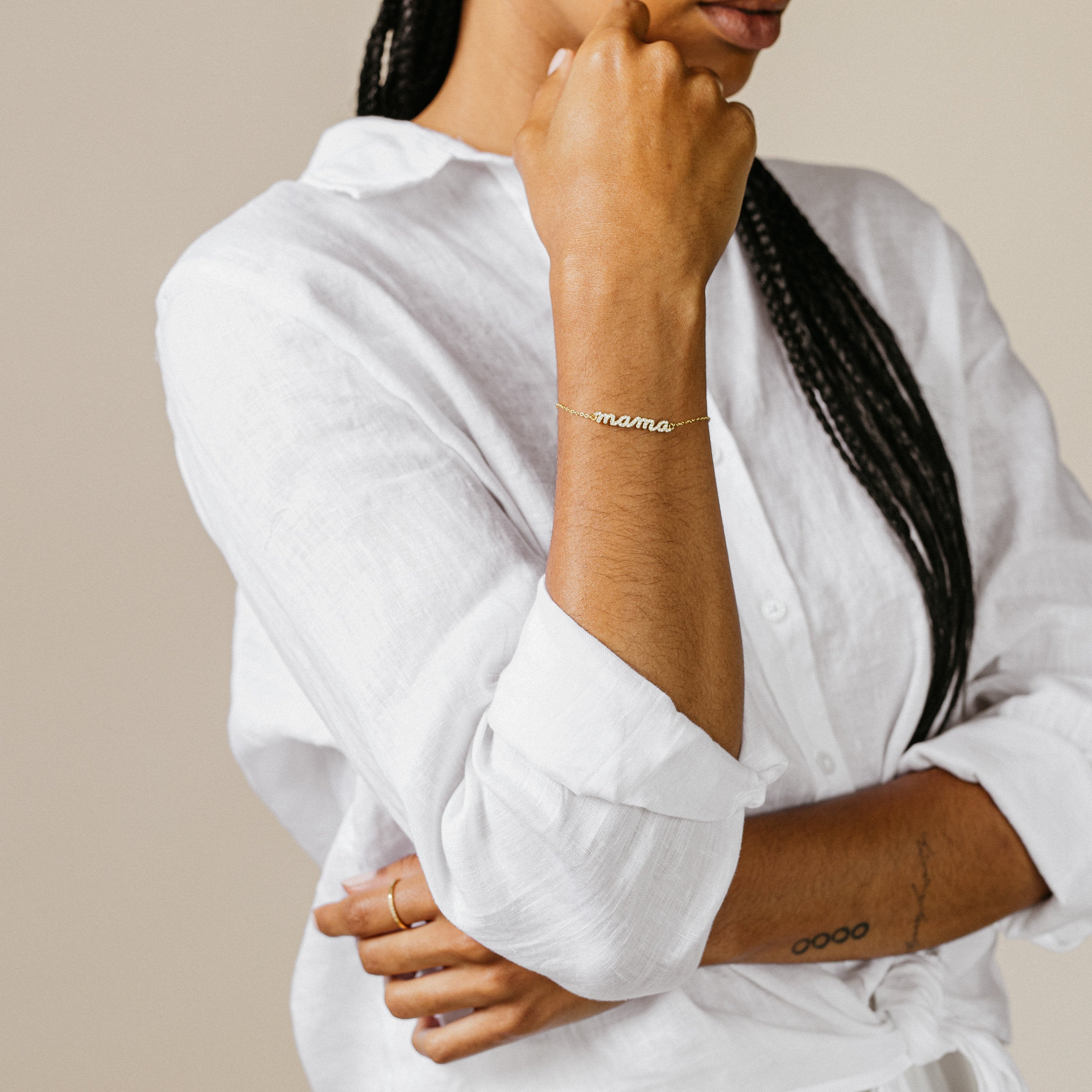 A person in a white shirt with braided hair celebrates stylish mama jewelry, wearing the Pave Mama Script Bracelet and a simple ring.