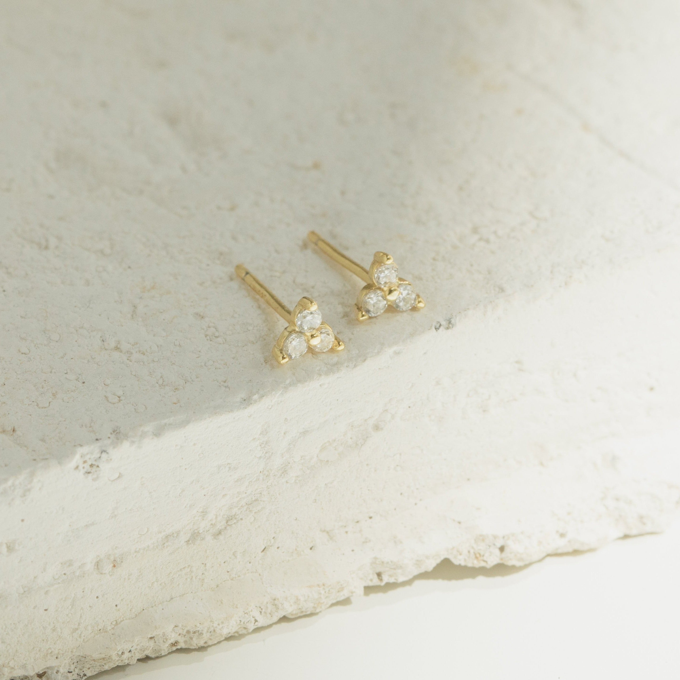 Mini Lily Studs: Dainty gold earrings each featuring three clear gemstones, ideal for everyday wear or stacking in cartilage piercings. Shown on a textured white stone surface.