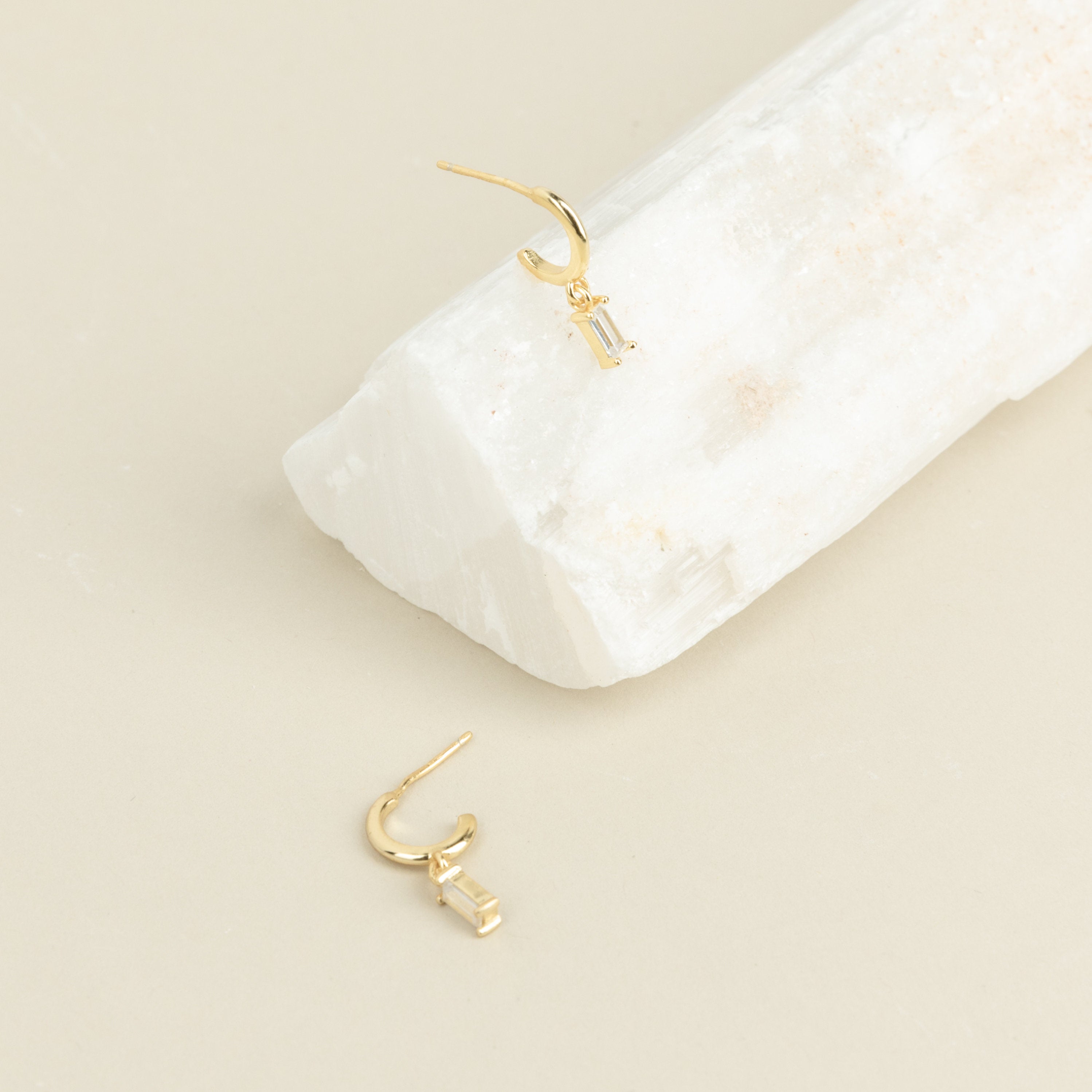 Two Lys Baguette Hoops, each featuring rectangular clear gemstones, display classic elegance—one rests on a white stone, the other on a beige surface.