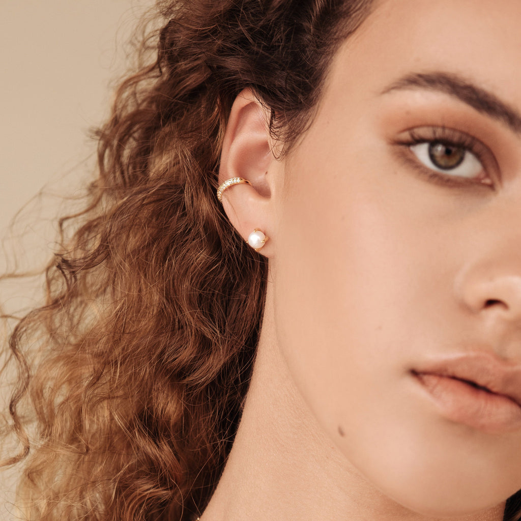 Close-up of a woman's face with curly hair, a gold hoop earring, and glimmering Mia Pave Cuffs diamond huggie earrings.