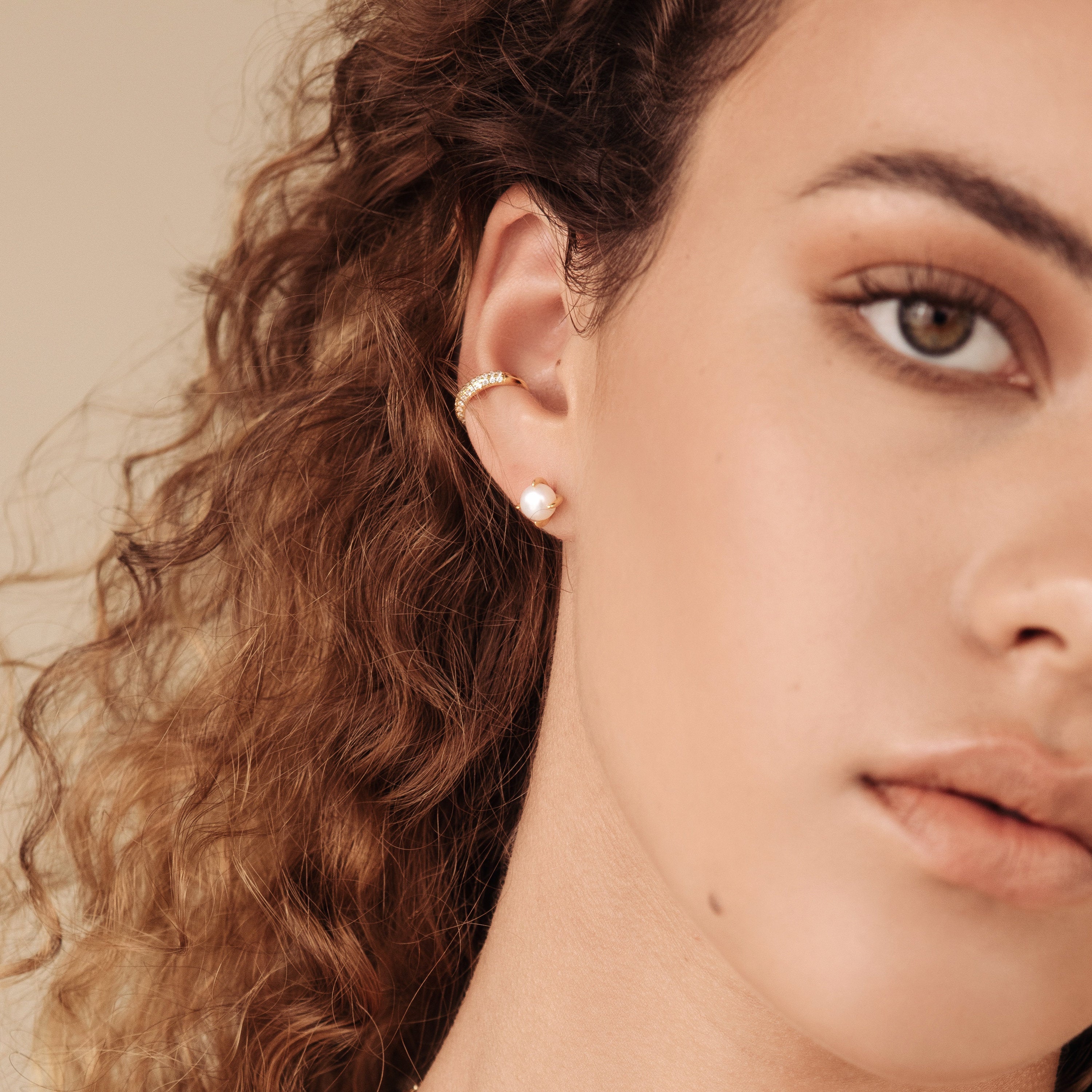 Close-up of a woman's face with curly hair, a gold hoop earring, and glimmering Mia Pave Cuffs diamond huggie earrings.