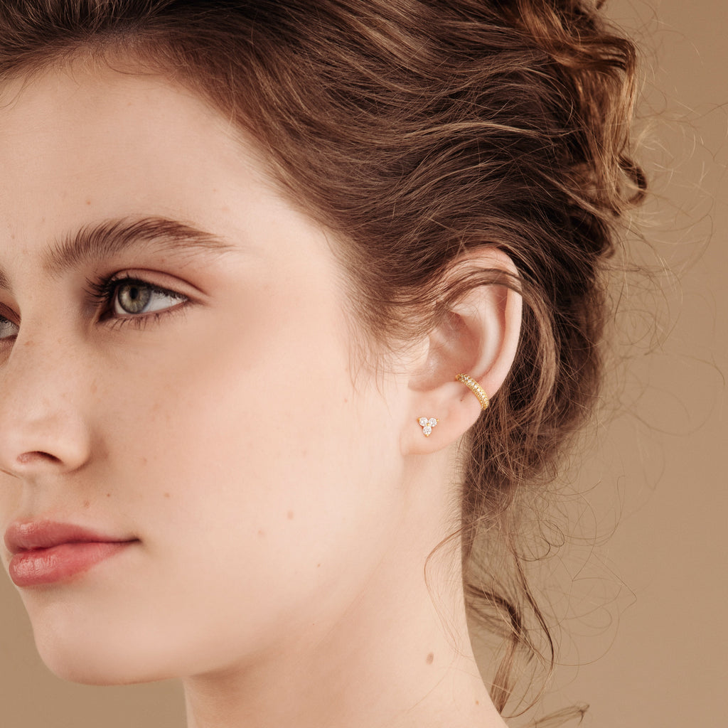 A woman with light skin and wavy brown hair pulled back wears Mia Pave Cuffs diamond huggie earrings against a beige background.