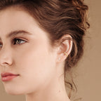 A woman with light skin and wavy brown hair pulled back wears Mia Pave Cuffs diamond huggie earrings against a beige background.