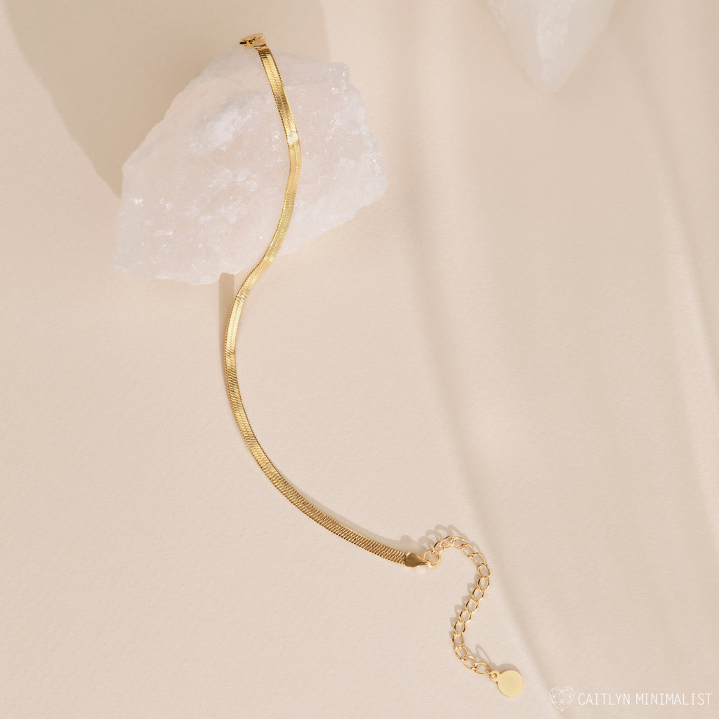 A Herringbone Anklet draped over a white crystal on a beige surface, highlighting the polished ankle jewelry's effortless elegance.