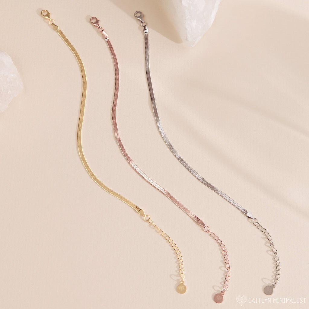 Three minimalist chain bracelets in gold, rose gold, and silver are displayed on a light beige background, perfectly complementing any polished ankle jewelry or pairing seamlessly with the Herringbone Anklet.