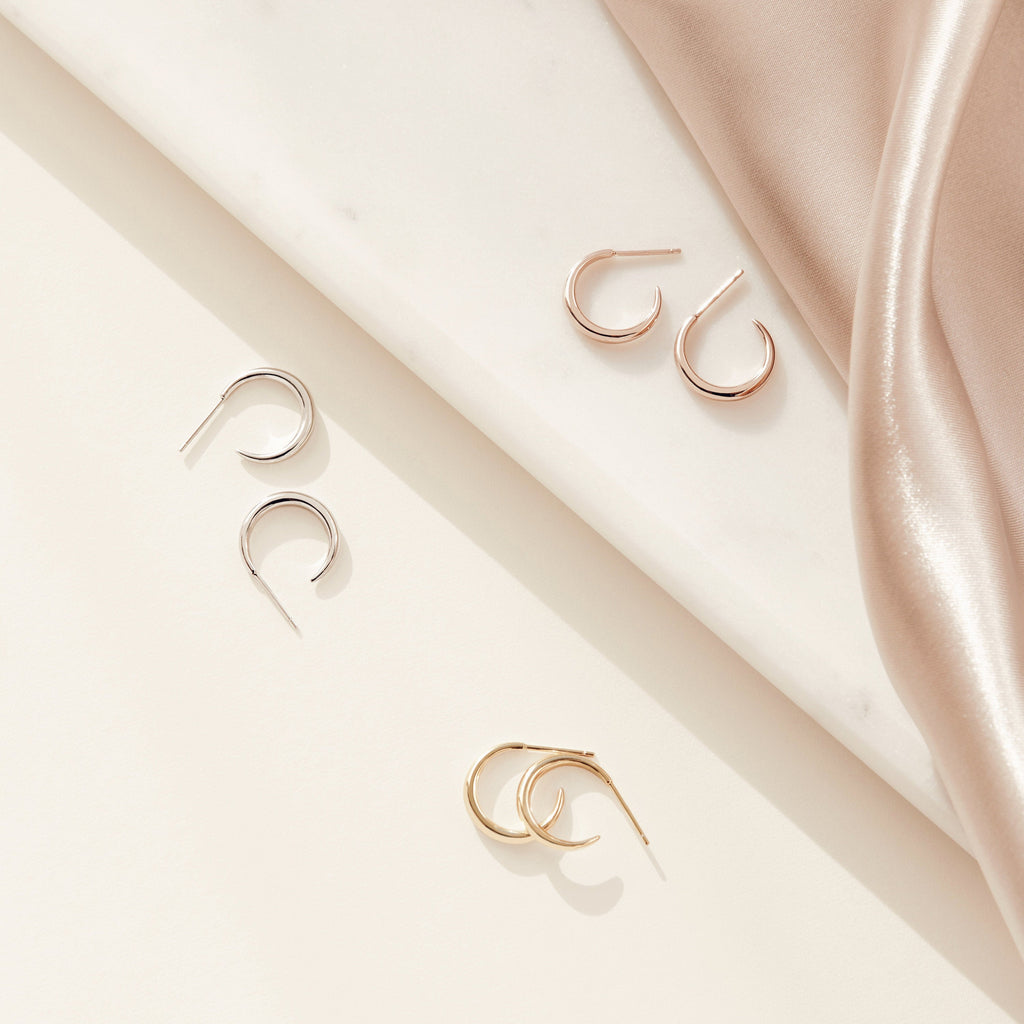 Three pairs of Chloe Hoops in silver, gold, and rose gold rest on marble and satin—polished bands that are ideal for everyday wear.
