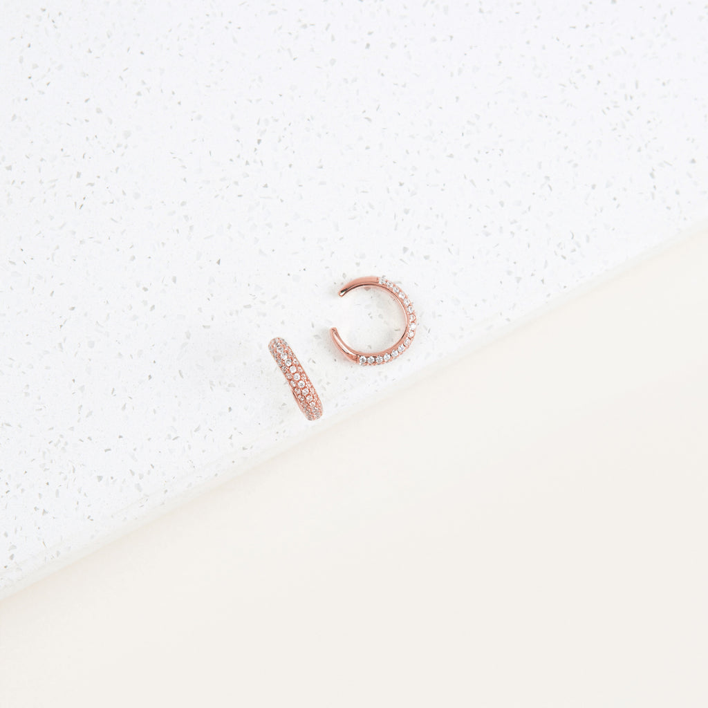 The Mia Pave Cuffs are rose gold earrings with small clear stones, sparkling beautifully on a white speckled surface.