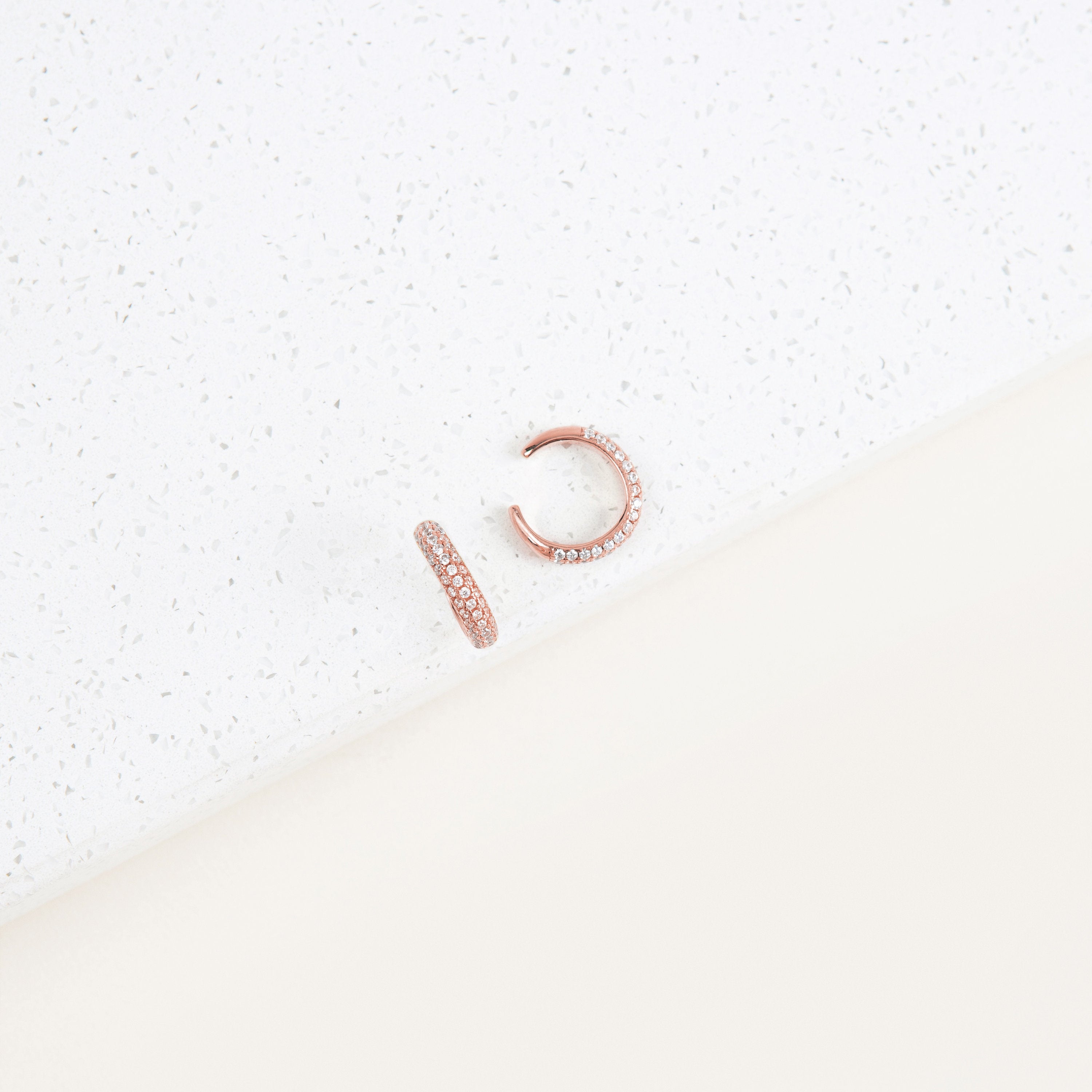 The Mia Pave Cuffs are rose gold earrings with small clear stones, sparkling beautifully on a white speckled surface.