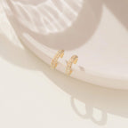 Two Mia Pave Cuffs adorned with small gold crystals are displayed on a pale, round ceramic surface under gentle sunlight.
