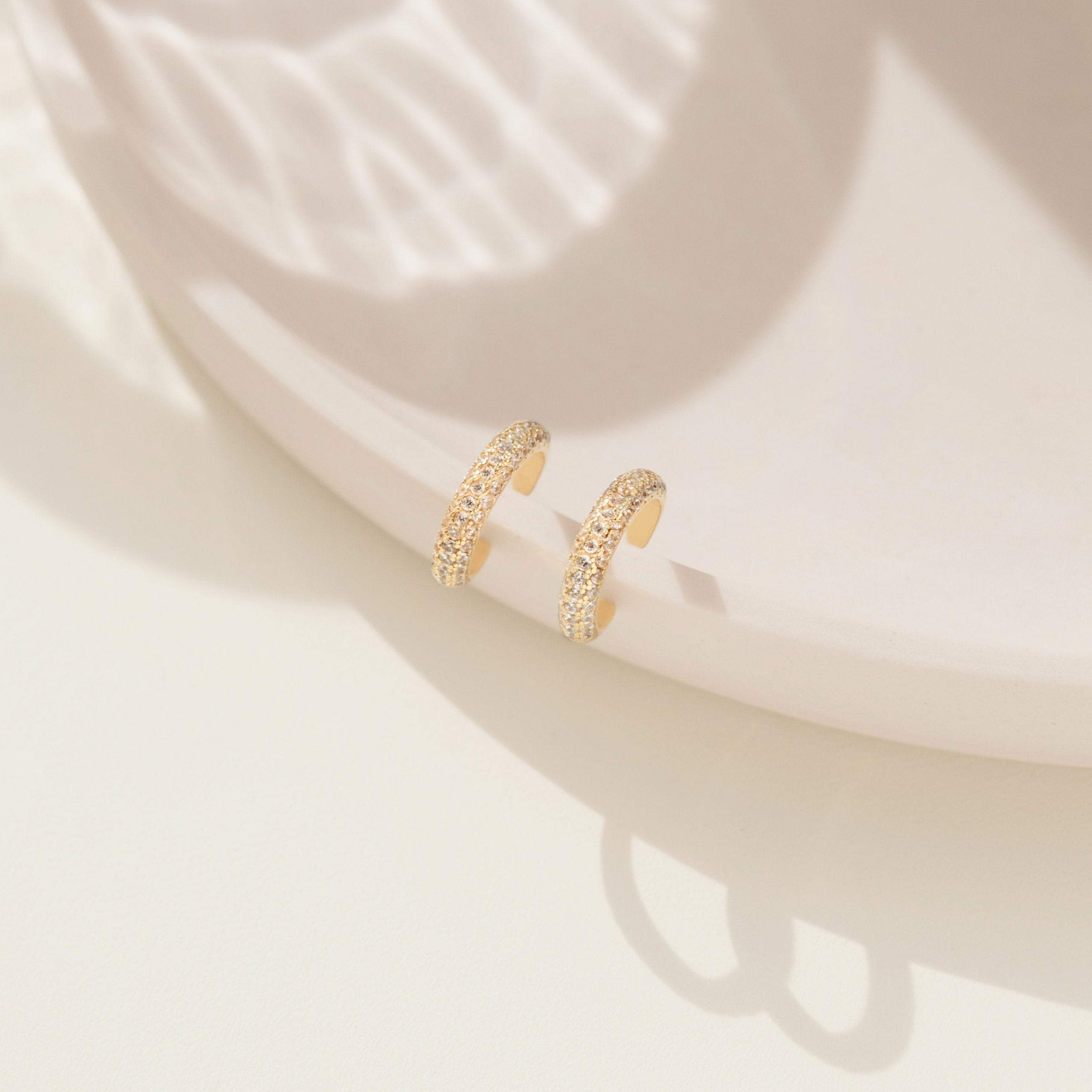 Two Mia Pave Cuffs adorned with small gold crystals are displayed on a pale, round ceramic surface under gentle sunlight.