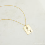 The Birth Flower Tag Necklace features a rectangular gold pendant engraved with a rose, displayed on a light textured surface—an ideal choice for personalized birth month jewelry.