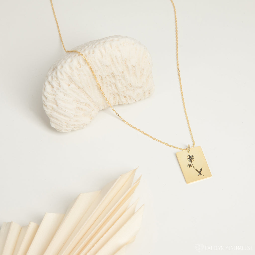 The Birth Flower Tag Necklace features an engraved floral design, shown on a white shell with soft fabric—an elegant personalized jewelry piece celebrating your birth month.