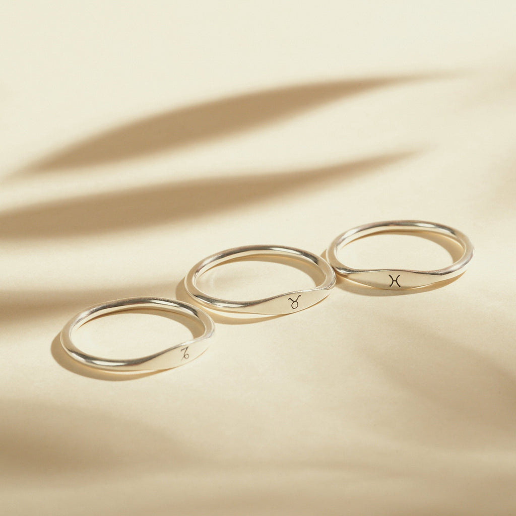 Three Skinny Zodiac Rings, each engraved with an astrological sign, are displayed in a row on a beige surface with soft shadows.