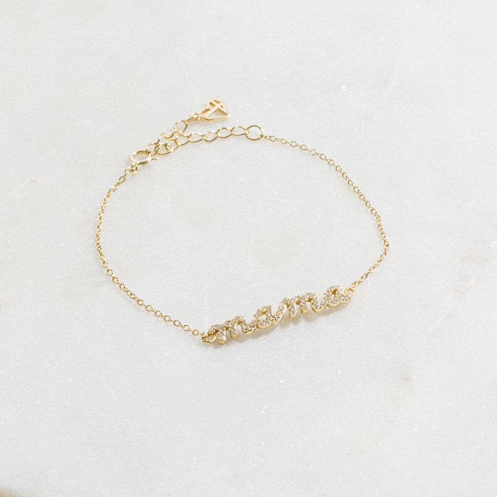 The Pave Mama Script Bracelet in gold, with "mama" spelled out in sparkling detail, rests on white marble—a stylish and elegant piece of jewelry perfect for celebrating motherhood.