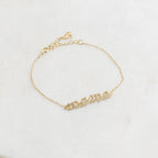 The Pave Mama Script Bracelet in gold, with "mama" spelled out in sparkling detail, rests on white marble—a stylish and elegant piece of jewelry perfect for celebrating motherhood.